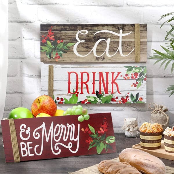 Three decorative Wooden Christmas Signs read "Eat," "Drink," and "Be Merry," displayed on a table with fruit, bread, muffins, and a rustic kitchen background.