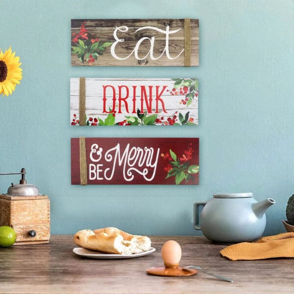 Three Wooden Christmas Signs on a blue wall read "Eat," "Drink," and "Be Merry." Below, a teapot, cup, bread, and coffee grinder rest on a wooden table, creating a festive scene.