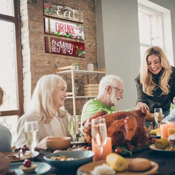 A group of people sit around a table enjoying a meal together, with festive Wooden Christmas Signs reading "Eat," "Drink," and "Be Merry" decorating the wall behind them.