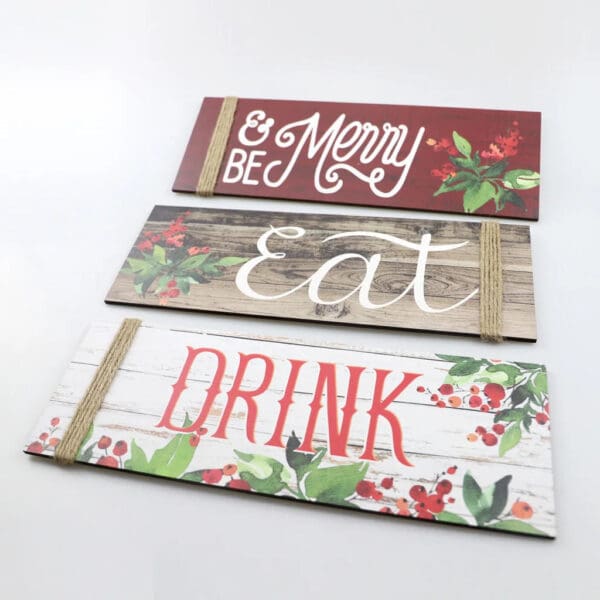 Three decorative Wooden Christmas Signs feature "Be Merry" on red, "Eat" on wood grain, and "Drink" on white, each adorned with holiday-themed foliage and twine accents.