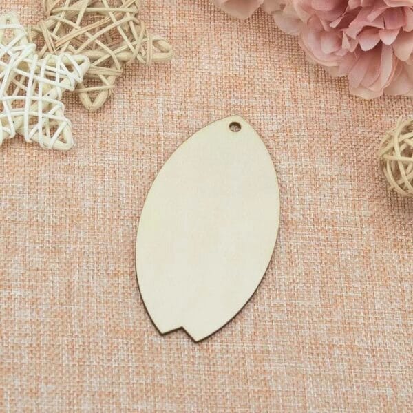 A plain, oval-shaped Wood Surfboard Cutout with a small hole near one end is placed on a textured pink fabric alongside rattan ornaments and pink flowers.
