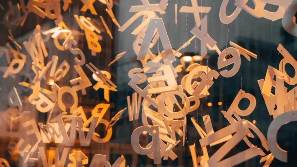 Numerous suspended cut-out letters in various fonts and orientations hang in front of a glass window, offering inspiring wooden letter decoration ideas with their abstract composition.