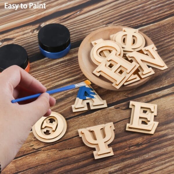 A hand paints a wooden letter "A" blue with a brush. Several unpainted wooden Greek letters and two paint jars rest on a wooden surface.
