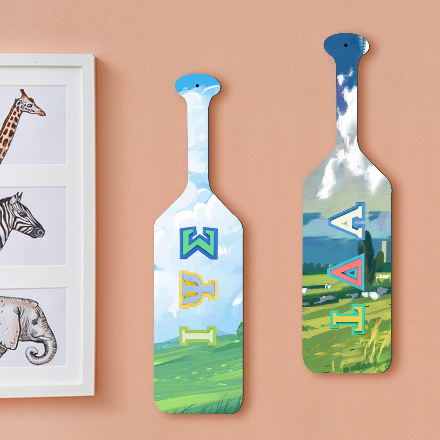 Two painted wooden paddles with blue skies and wooden Greek letters hang on a peach wall next to a framed animal illustration featuring a giraffe, zebra, and elephant.
