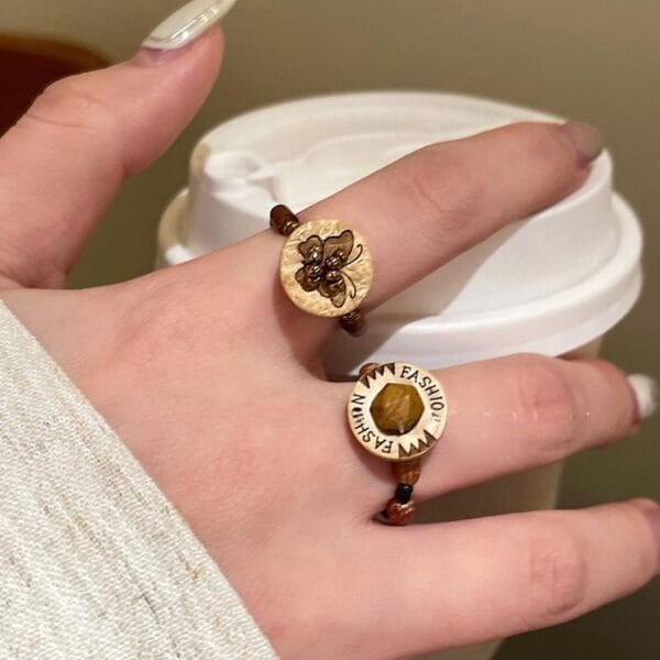 A hand holding a white takeaway coffee cup, adorned with eco-friendly wood jewelry—two brown and gold rings with unique decorative designs on the index and middle fingers.