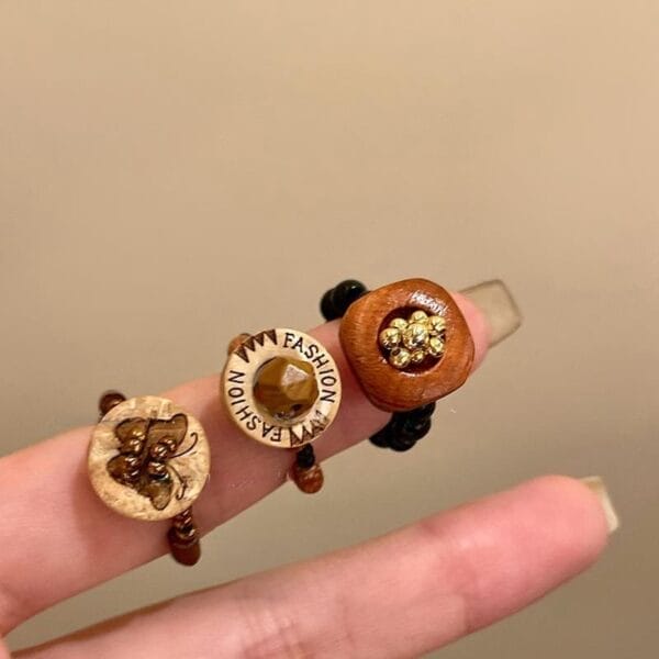 Three beaded rings with circular wooden and decorative centerpieces, showcasing eco-friendly wood jewelry, are worn on two fingers against a plain background.