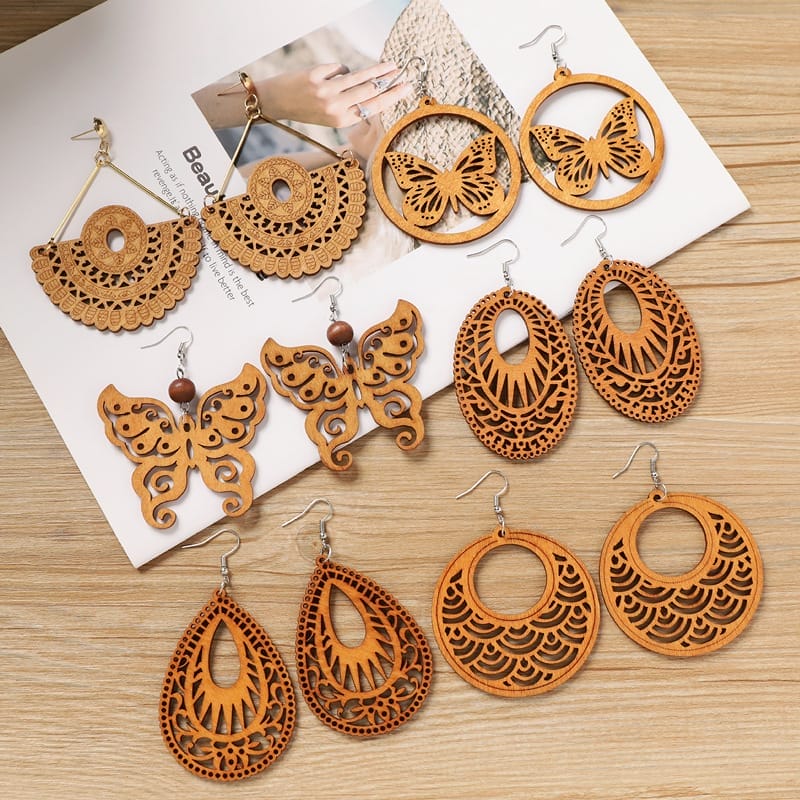 Five pairs of intricately carved wooden earrings in various shapes, including butterflies, circles, and teardrops, displayed on a wooden surface with a magazine in the background.