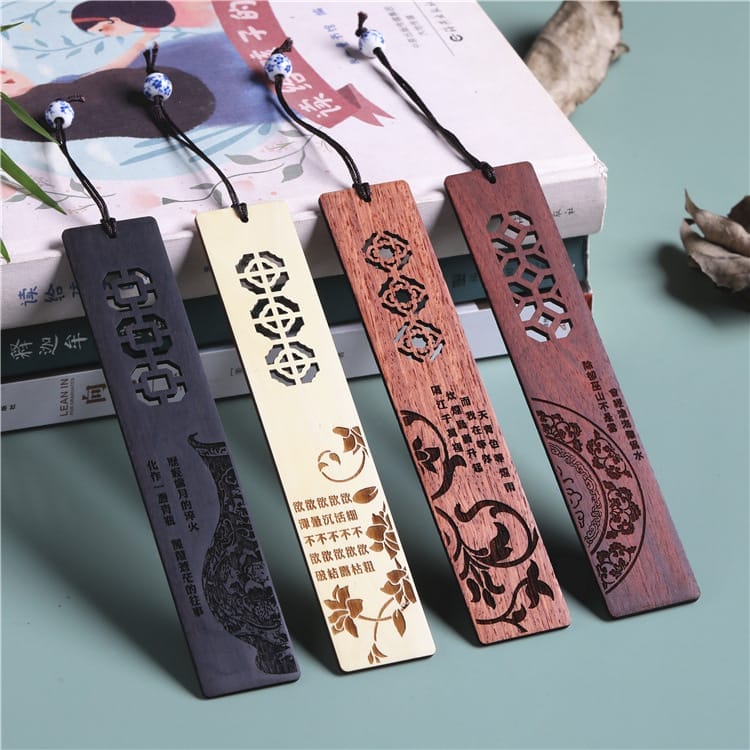 Four wooden bookmarks with intricate cut-out and engraved designs are displayed on top of books, each with a decorative string and bead attached.