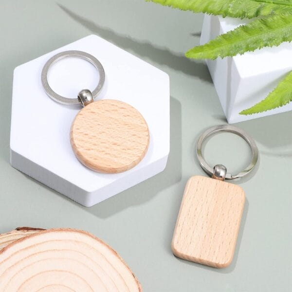 Two plain wooden keychains, one round and one rectangular, are displayed on a light surface with decorative white shapes and green leaves.