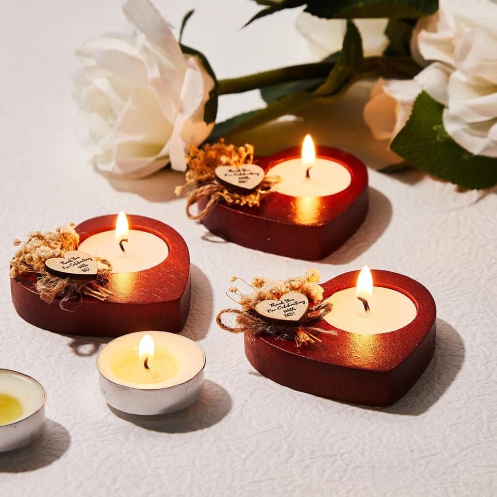 Three heart-shaped candles and two tealights are lit on a white surface, with white artificial flowers in the background. Two heart candles have small decorative tags.