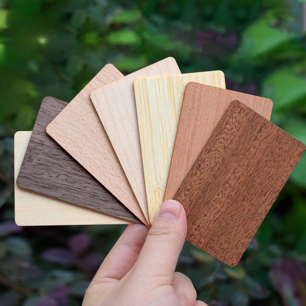 A hand holds a fanned-out set of seven rectangular wood veneer samples in various shades, against a blurred green outdoor background.