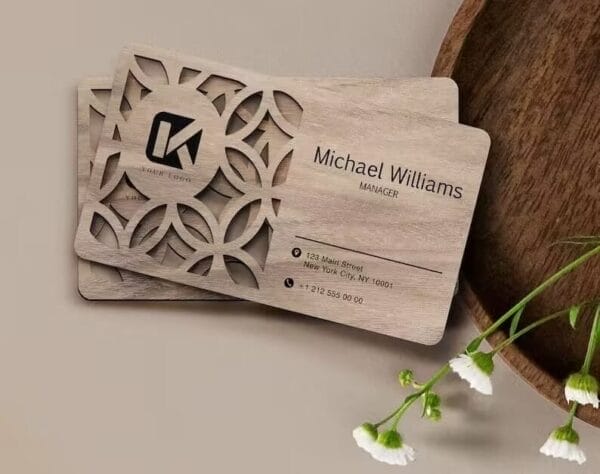 Two Laser Cut Business Cards with a floral cutout on the left display Michael Williams, Manager, along with contact details and social icons. These Custom Wooden Business Cards are placed beside white flowers.