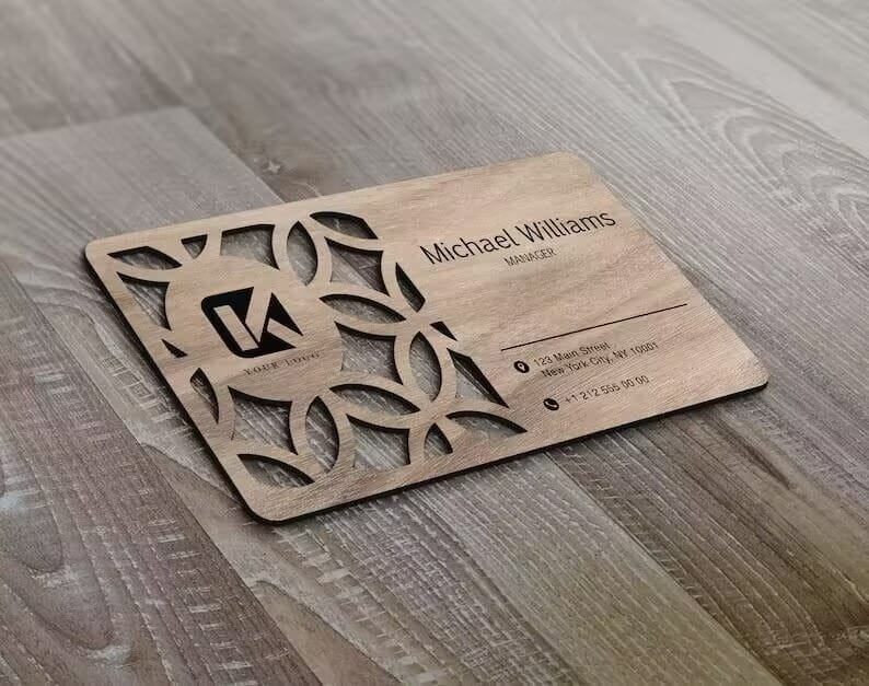A Custom Wooden Business Card with geometric cutout designs and printed contact details for Michael L. Williams, Manager, is displayed on a wooden surface.