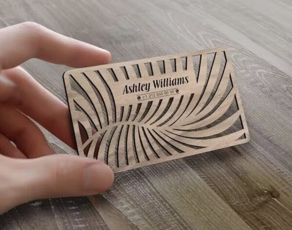A hand holds a personalized wood card with intricate leaf-like cutouts, displaying the name "Ashley Williams" and a phone number.