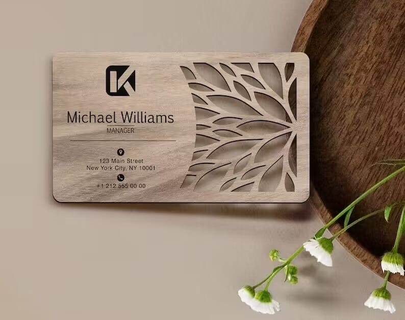 A custom wooden business card for Michael Williams, Manager, featuring a leaf pattern cutout, contact details, and placed next to a wooden tray and white flowers—an elegant example of personalized wood cards.