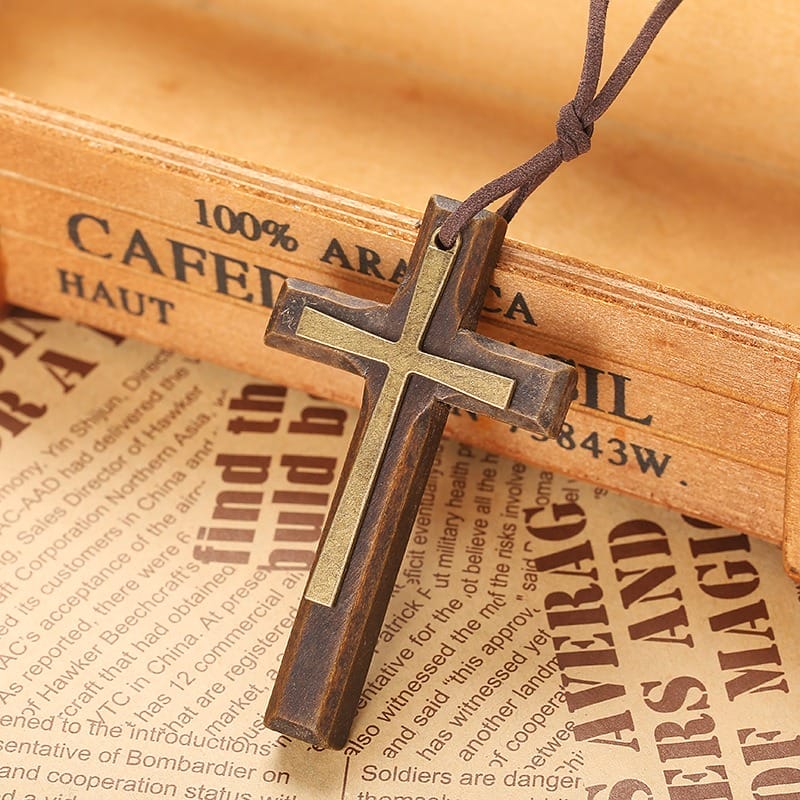 A wooden cross necklace with a metal inlay hangs on a brown cord, resting on a vintage book and newspaper background—a timeless vintage necklace perfect as a unisex sweater chain accessory.