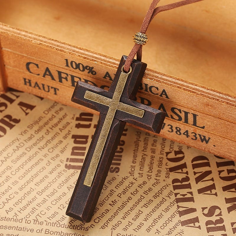 A vintage necklace featuring a wooden cross pendant with a metallic accent on a brown leather cord, displayed on printed paper and a wooden box background.