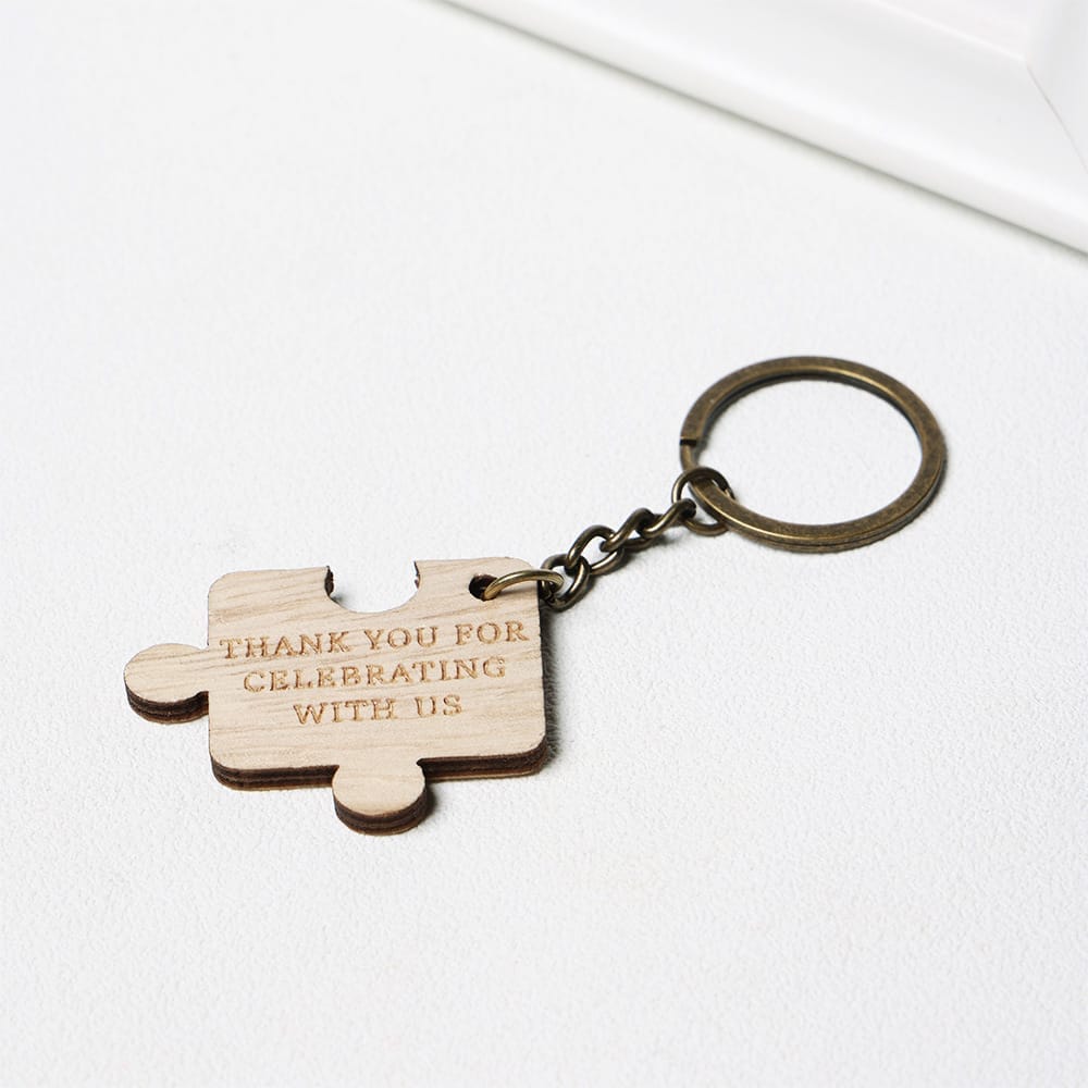 A metal keyring with a wooden puzzle piece charm engraved with the words "Thank you for celebrating with us" lies on a white surface.