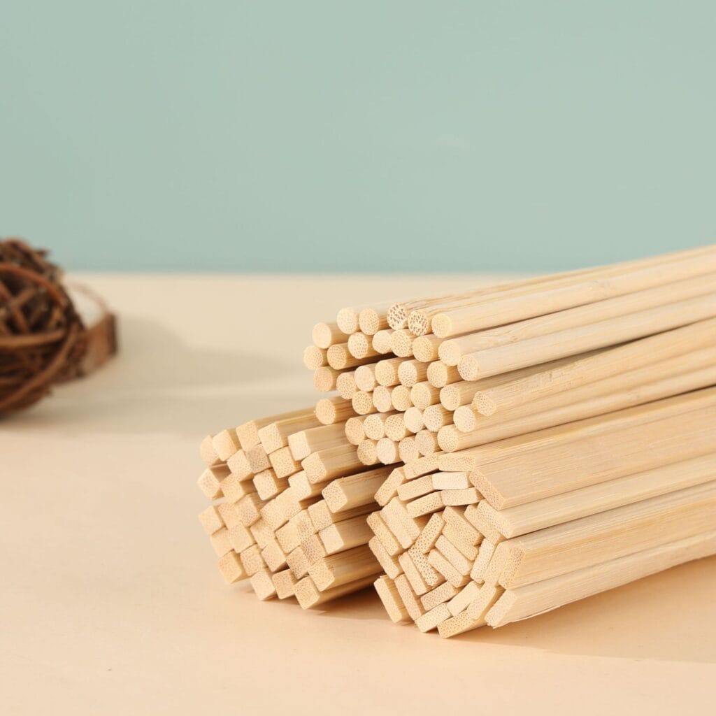 Bundles of light-colored chopsticks, resembling wooden sticks for crafts, some with round ends and some with square ends, are stacked on a beige surface against a light blue background.