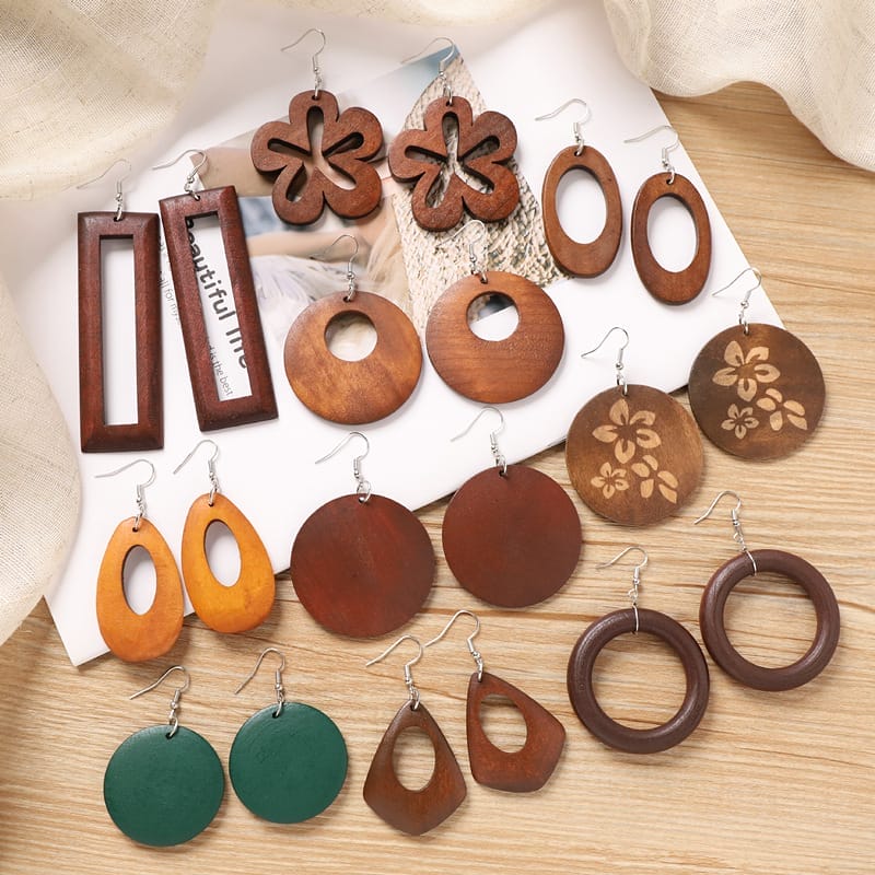 A collection of various wooden and green painted earrings in different shapes and designs, displayed on a wooden surface with fabric and a magazine.