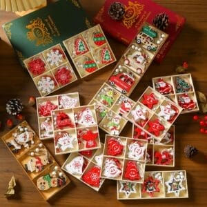 Assorted Christmas tree ornaments in festive shapes and colors are arranged in segmented boxes on a wooden table with books, pine cones, and decorative berries nearby.