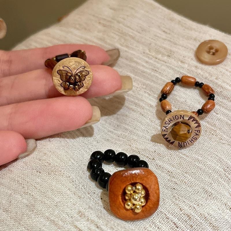 A hand displays a wooden ring with a butterfly design; two other beaded rings with wooden accents and text sit on a beige fabric surface nearby.