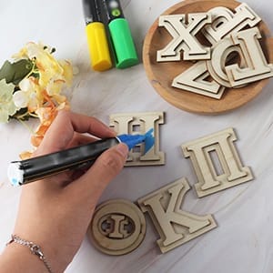 A hand colors wooden Greek letters with a blue marker, while more wooden Greek letters and colorful markers are scattered on a white surface.