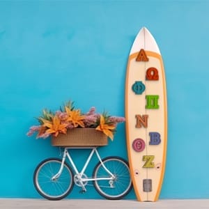 A white surfboard with colorful wooden Greek letters leans against a blue wall beside a white bicycle with a basket of orange and purple flowers.