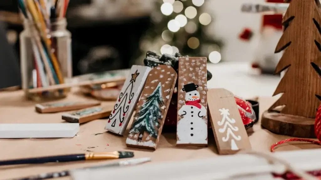 Wooden crafts painted with Christmas designs, like a tree and a snowman, are displayed on a table with paintbrushes and supplies—perfect DIY Christmas wood cutouts for festive home decor. A blurred Christmas tree glows in the background.
