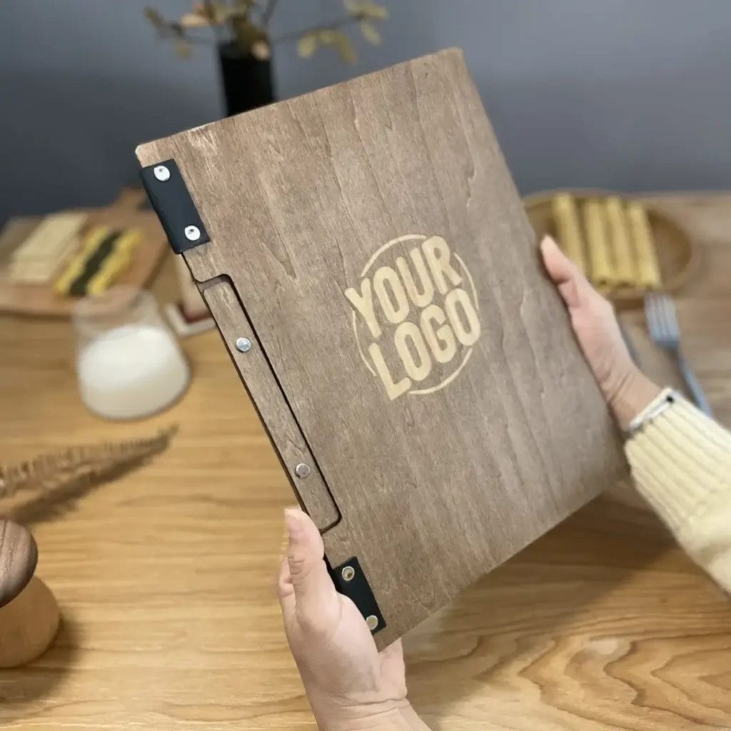 A person holds a wooden menu with metal hinges and a circular "YOUR LOGO" design on the front, sitting at a wooden table set for a meal.