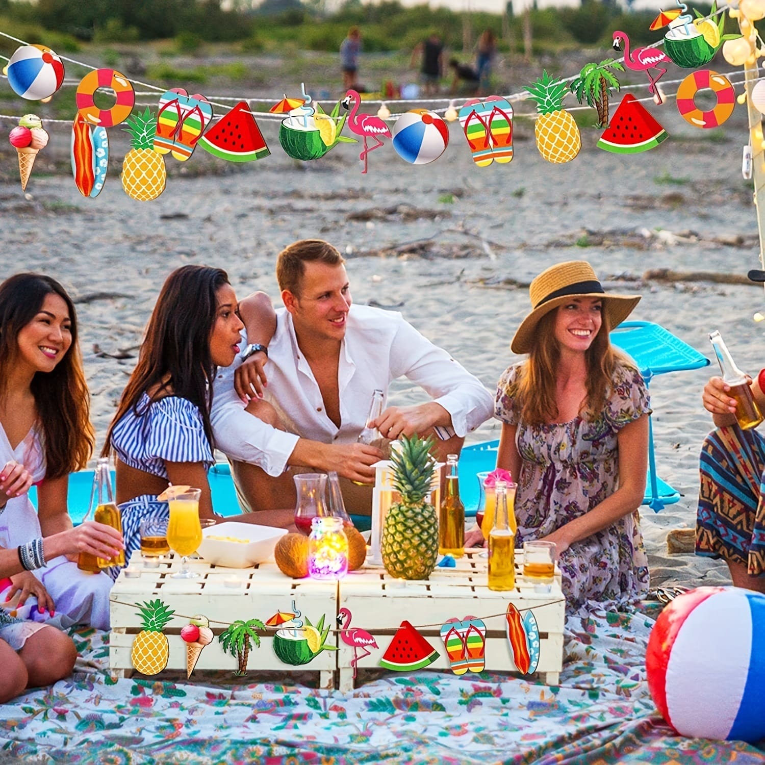 A group of people sit on a Hawaiian Beach enjoying drinks and food, surrounded by colorful tropical decorations, a picnic setup, and a playful watermelon pendant nestled among the vibrant decor.