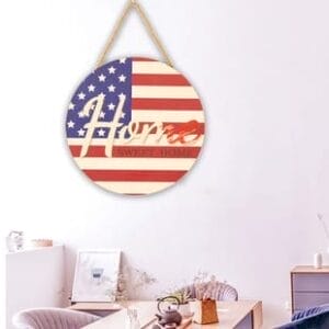 A round wooden American flag sign with "Home Sweet Home" text hangs on a light-colored wall above a desk adorned with plants and small decor items.