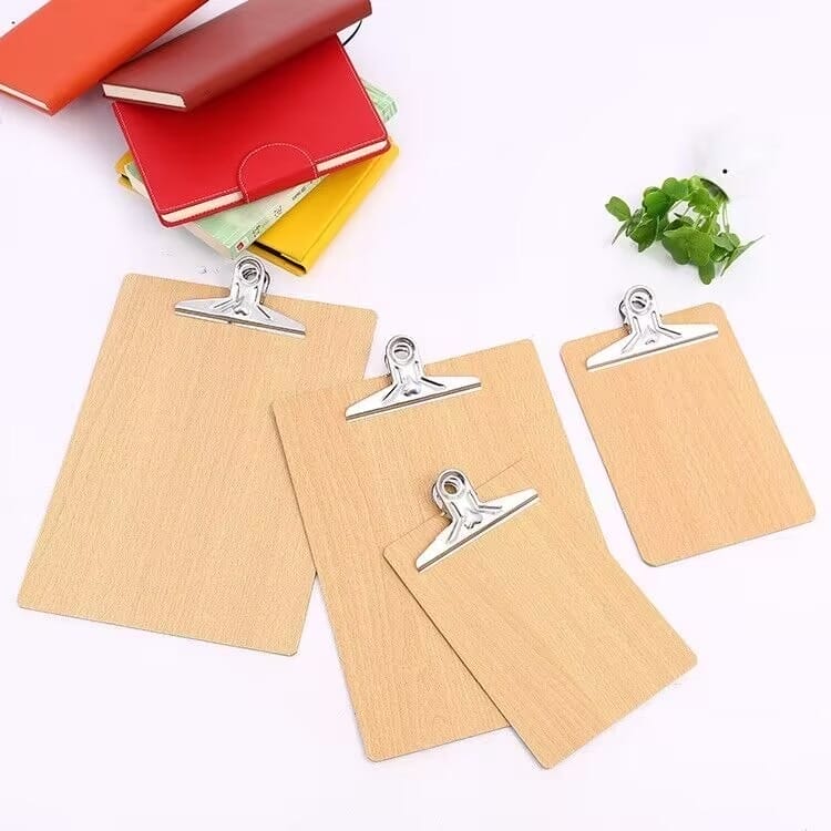 Four wooden clipboards of different sizes, including A4 clipboards, are arranged on a white surface, with books and a small green plant in the background. Perfect as wholesale wooden clipboards or chic butterfly clip menu holders.