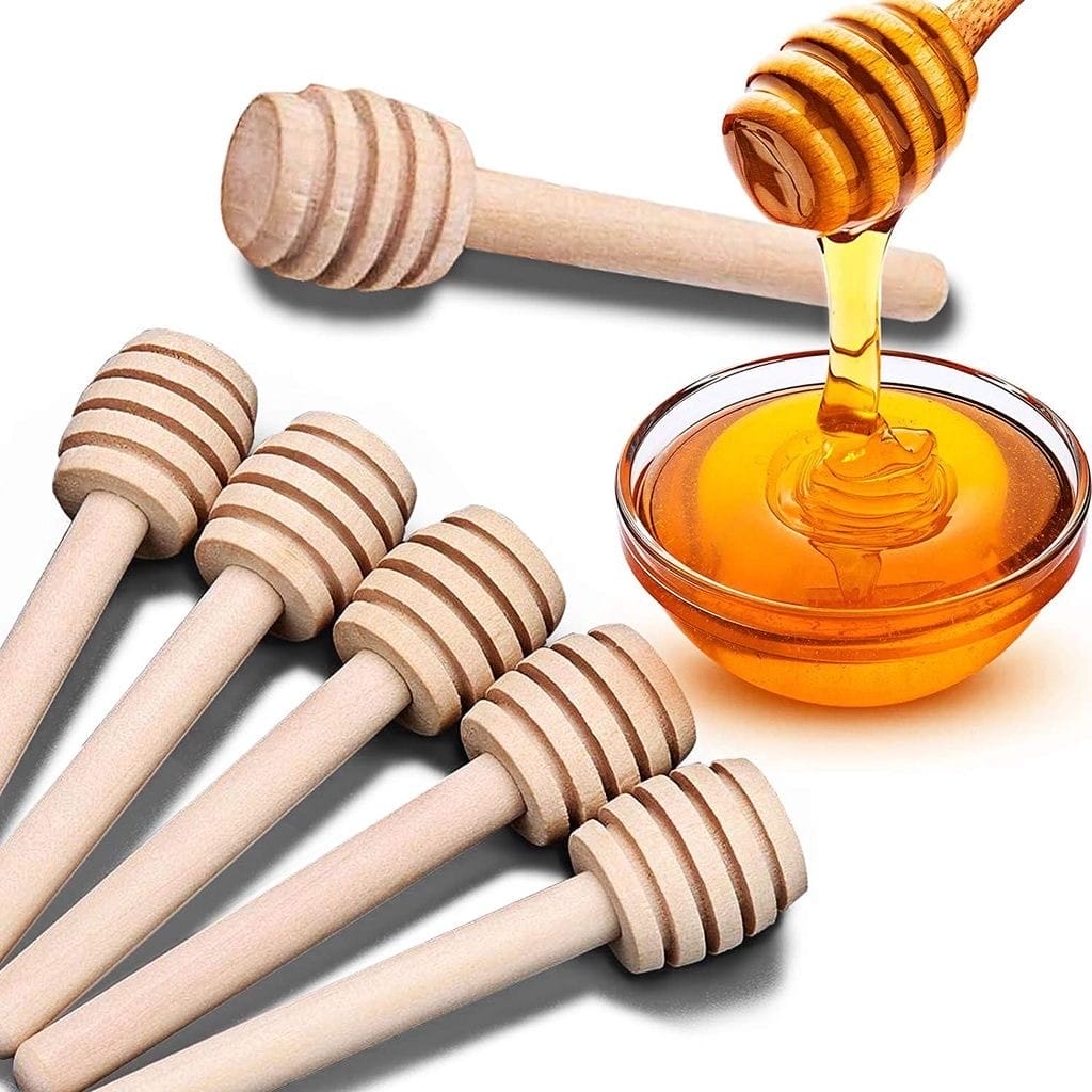 Six wooden honey dippers are arranged on a white surface next to a glass bowl filled with honey, with one dipper drizzling honey into the bowl.