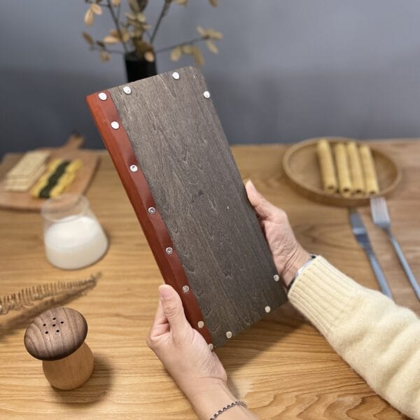 Hands holding a dark wood menu cover with a brown leather spine and silver rivets. Wholesale restaurant menu.