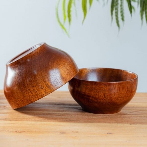 Two polished wooden salad bowls, one upright and one tilted on its side, are placed on a light wooden surface with green plant leaves in the background. These durable wood bowls add a natural touch to any setting.