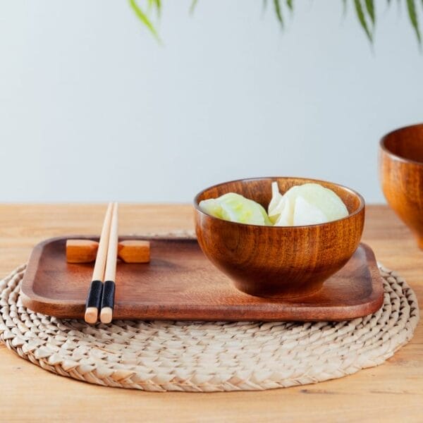 A wooden bowl with sliced vegetables on a wooden tray, chopsticks on a holder, and a woven placemat on a wooden table—perfect for showcasing Small Wood Bowls in any setting.