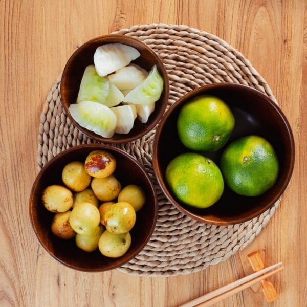 Three wood bowls on a woven mat contain green citrus fruits, yellow round fruits, and white dumplings; a pair of orange chopsticks rests nearby on a wooden surface. Perfect for wholesale or as elegant wooden salad bowls.
