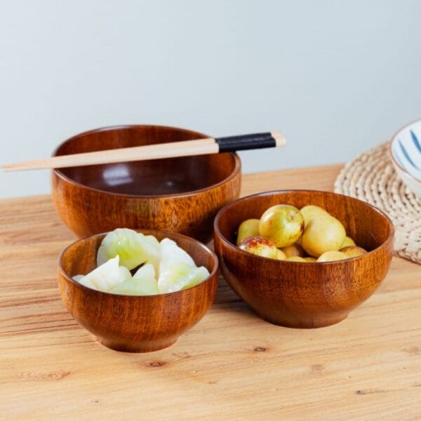 Three wooden salad bowls on a wooden table—one filled with apples, another with white dumplings, and one empty—with chopsticks resting on the largest bowl.