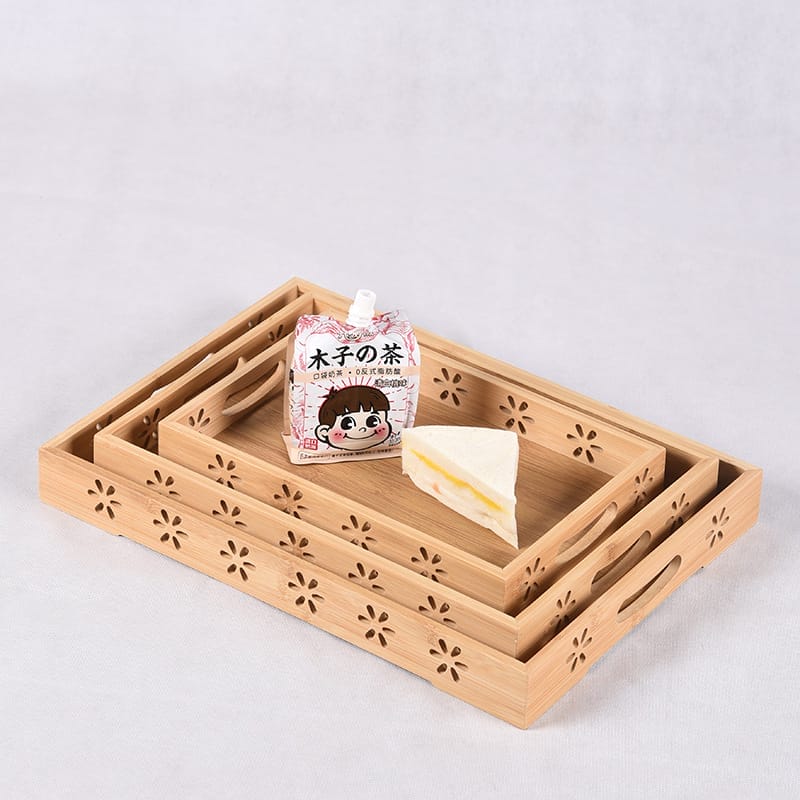 Three nested wooden trays with floral cutout patterns, holding a packaged drink and a slice of cake, displayed on a white surface.