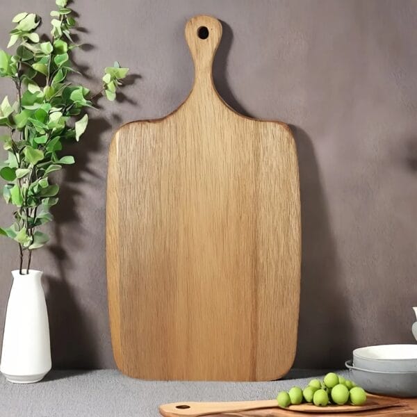 A large acacia wood cutting board with a handle stands upright on a surface. Nearby, a vase with green leaves, a small bowl of green olives, and a wooden tray create an inviting charcuterie board setup.