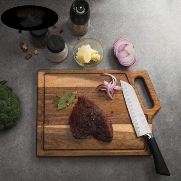 A raw steak with spices and bay leaves on an Acacia Wood Cutting Board, a knife, onion, butter, salt, pepper, and broccoli on a gray countertop.