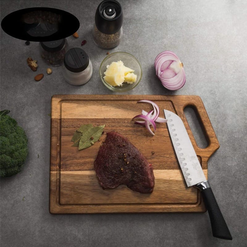 A raw steak with spices and bay leaves on an Acacia Wood Cutting Board, a knife, onion, butter, salt, pepper, and broccoli on a gray countertop.