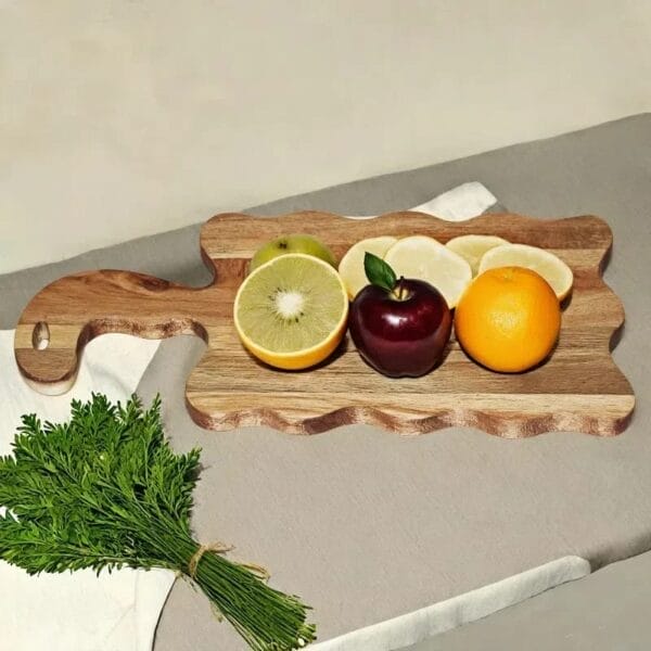 A round acacia wood cutting board with a green apple, kiwi, sliced lemon, red apple, and orange sits next to a bundle of leafy greens on a gray surface.