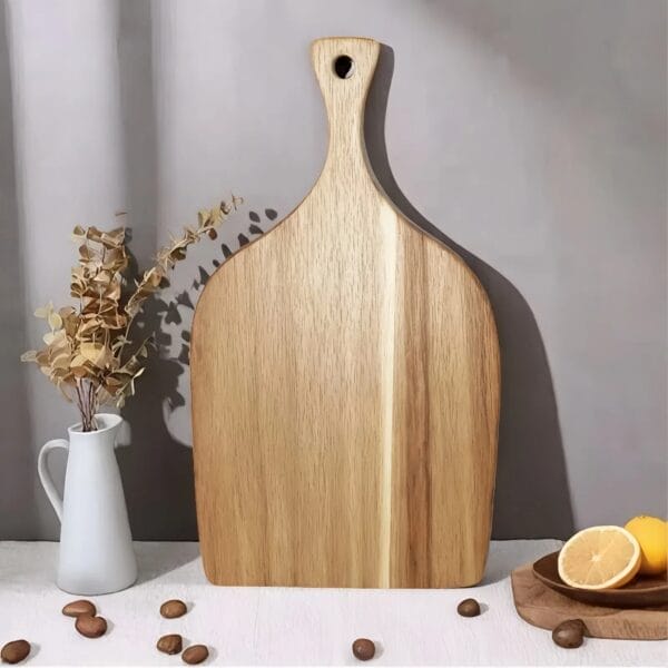 A round charcuterie board with a handle stands upright on a white surface beside a white vase with dried leaves, scattered nuts, and a halved lemon.