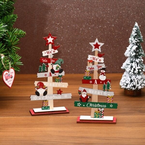Two small Christmas Wooden Ornaments with festive designs and text stand on a wooden surface beside a green artificial tree and a snowy miniature tree, making charming Christmas Tree Decorations.
