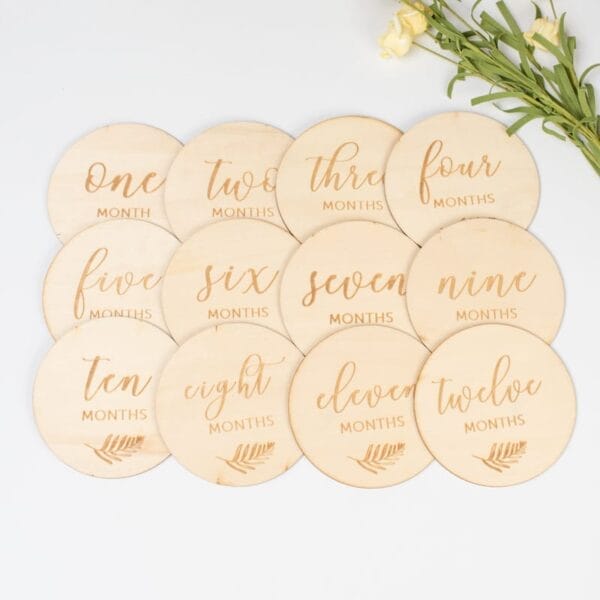 Twelve round wooden milestone cards, perfect as baby photo props, labeled one to twelve months and arranged in a grid with yellow flowers in the top right corner.