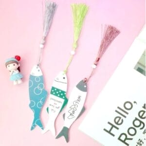 Three fish-shaped eco-friendly bookmarks with tassels are arranged on a pink surface next to a small doll figure and a partially visible book or magazine.