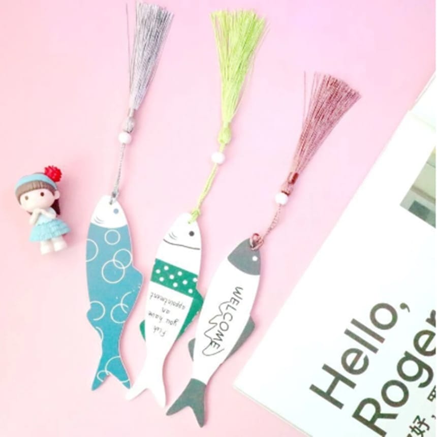 Three fish-shaped eco-friendly bookmarks with tassels are arranged on a pink surface next to a small doll figure and a partially visible book or magazine.