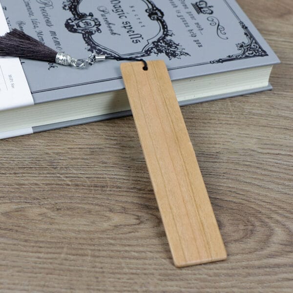 Wooden bookmark with tassel, resting on a book titled "Magic Spells". Custom standard size, engravable bookmark for distribution.